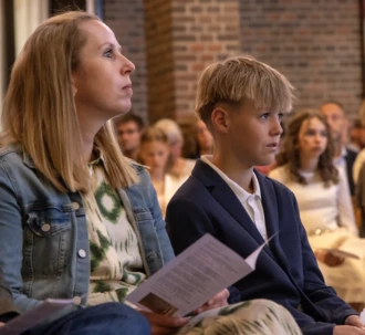En konfirmand og hans mor sidder i kirken og lytter opmærksomt under en konfirmation.
