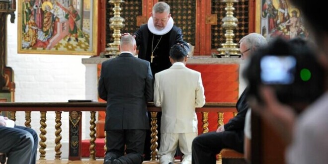 To mænd knæler foran alteret hvor en biskop velsigner parret i forbindelse med vielse