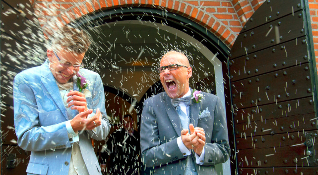 Billede af to nygifte mænd, der glade kommer ud af kirkedøren, mens gæsterne kaster ris på dem.