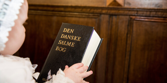 Dåbsbarn sidder hos en forælder på kirkebænken og kigger på en salmebog