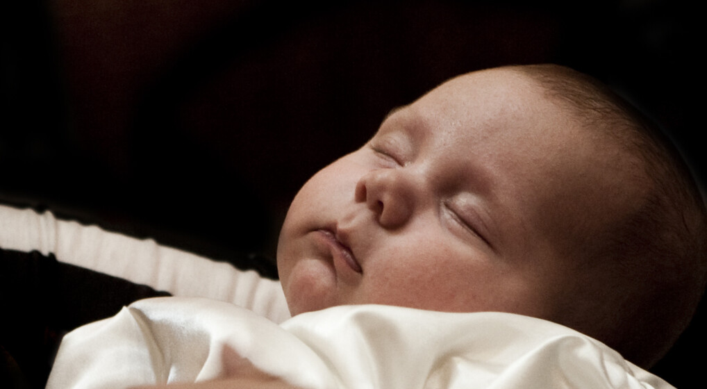 Baby i dåbskjole sover trygt i sin mors arm ved barnedåb