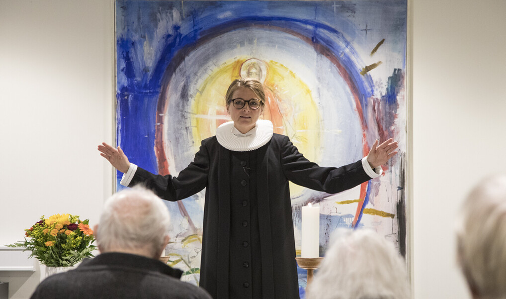 Præst i præstekjole foran alter løfter hænderne til velsignelse ved gudstjeneste i folkekirken