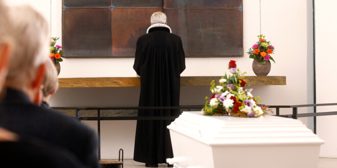 En hvid, blomsterpyntet kiste i et kirkerum med en præst i baggrunden.