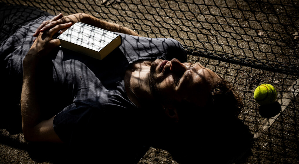 Billede af Mikkel Skovgaard, der ligger på ryggen på en tennisbane i skyggen af nettet med en salmebog på brystet