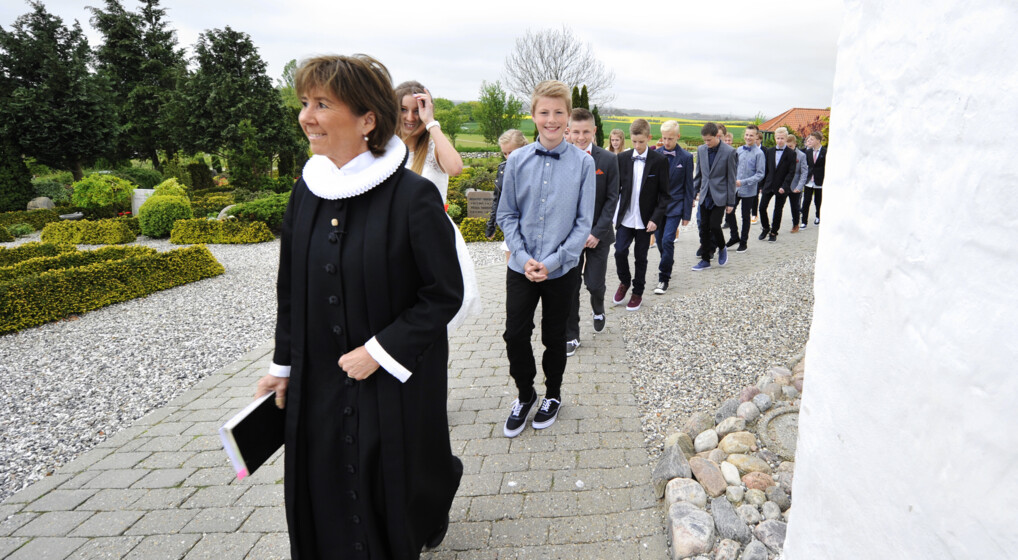 En præst går foran en lang række glade konfirmander uden for en kirke.